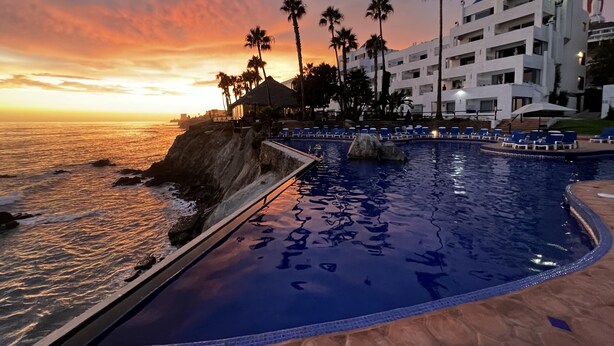 Une piscine d’un complexe hôtelier offrant une vue sur l’océan au coucher du soleil, des palmiers et un bâtiment moderne doté de balcons.
