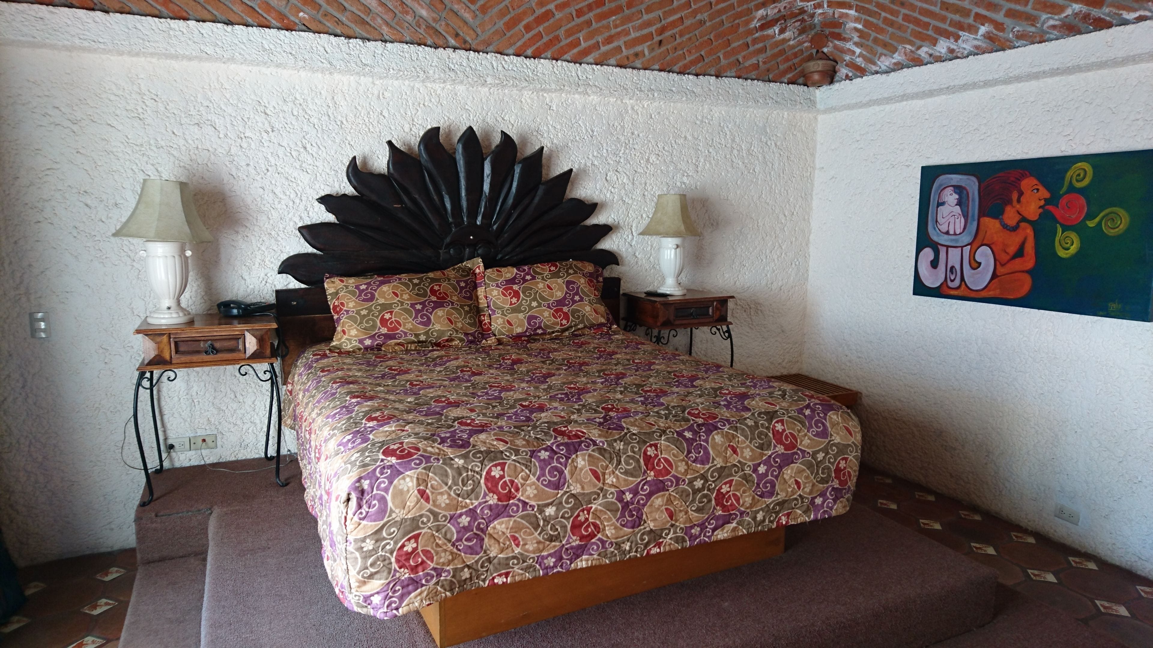 A bedroom with a large bed, two bedside tables, a decorative headboard, and a colorful painting on the wall.