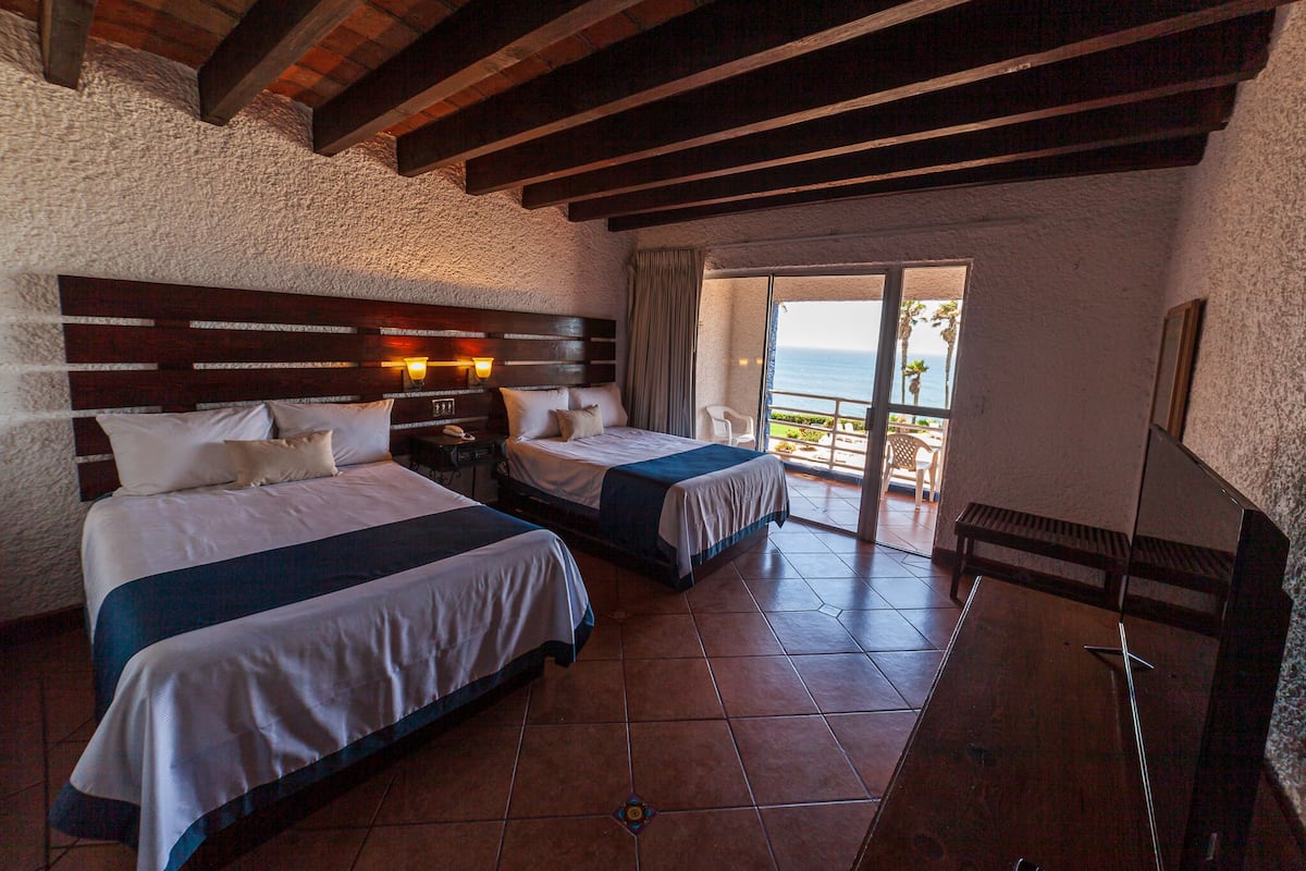 A hotel room with two beds, a wooden headboard, a nightstand, and a balcony with a view of the ocean.