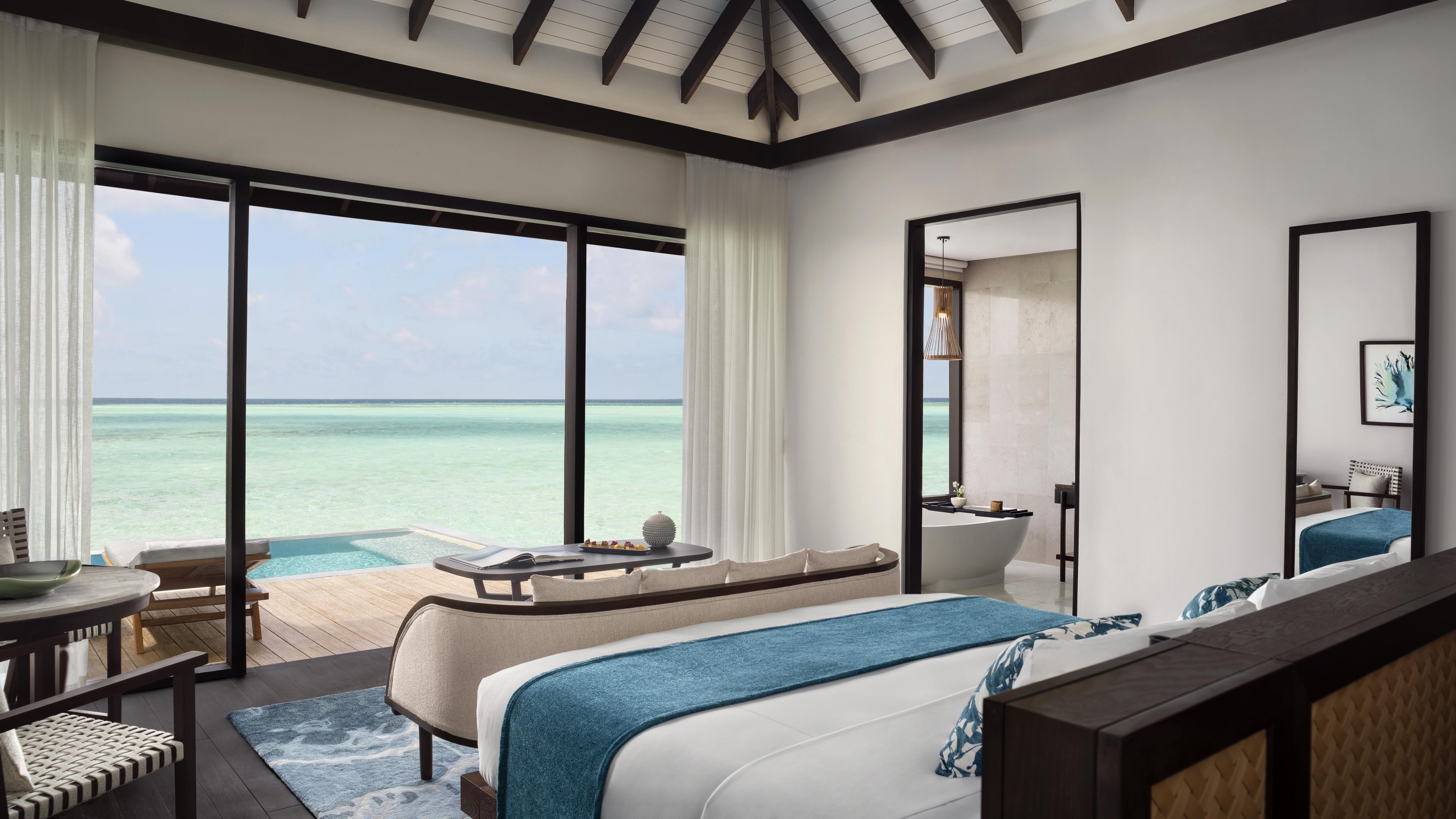 A modern bedroom with a large bed, a seating area, a small table with a book, and a view of the ocean.