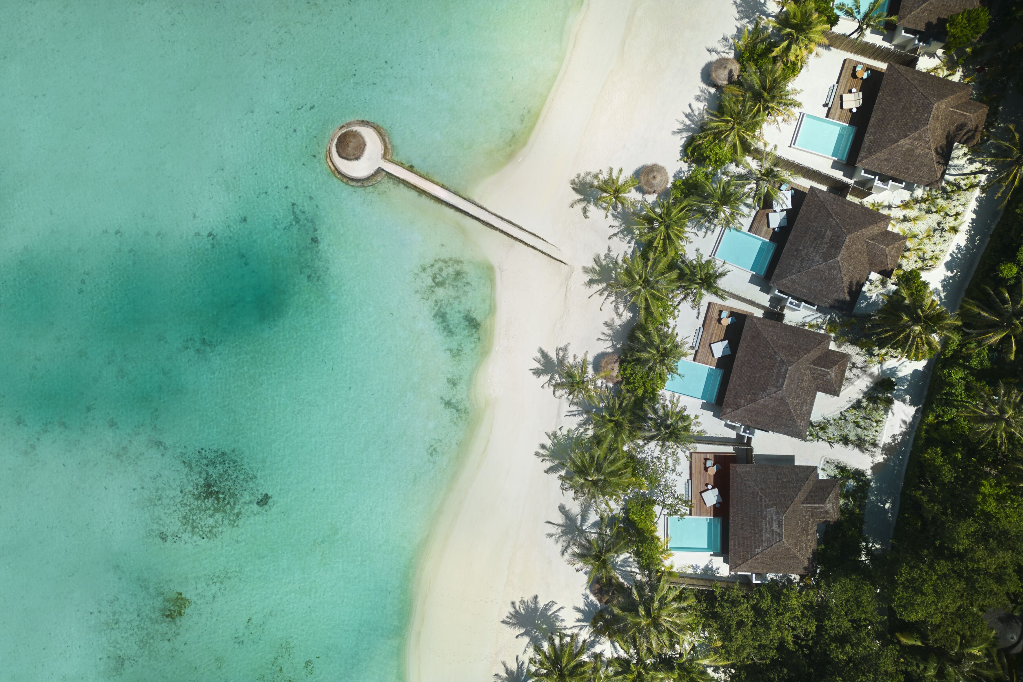 An aerial view of a beach resort with a swimming pool, thatched-roof huts, and palm trees.