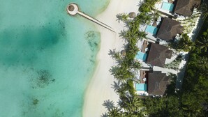 An aerial view of a beach resort with a swimming pool, thatched-roof huts, and palm trees.