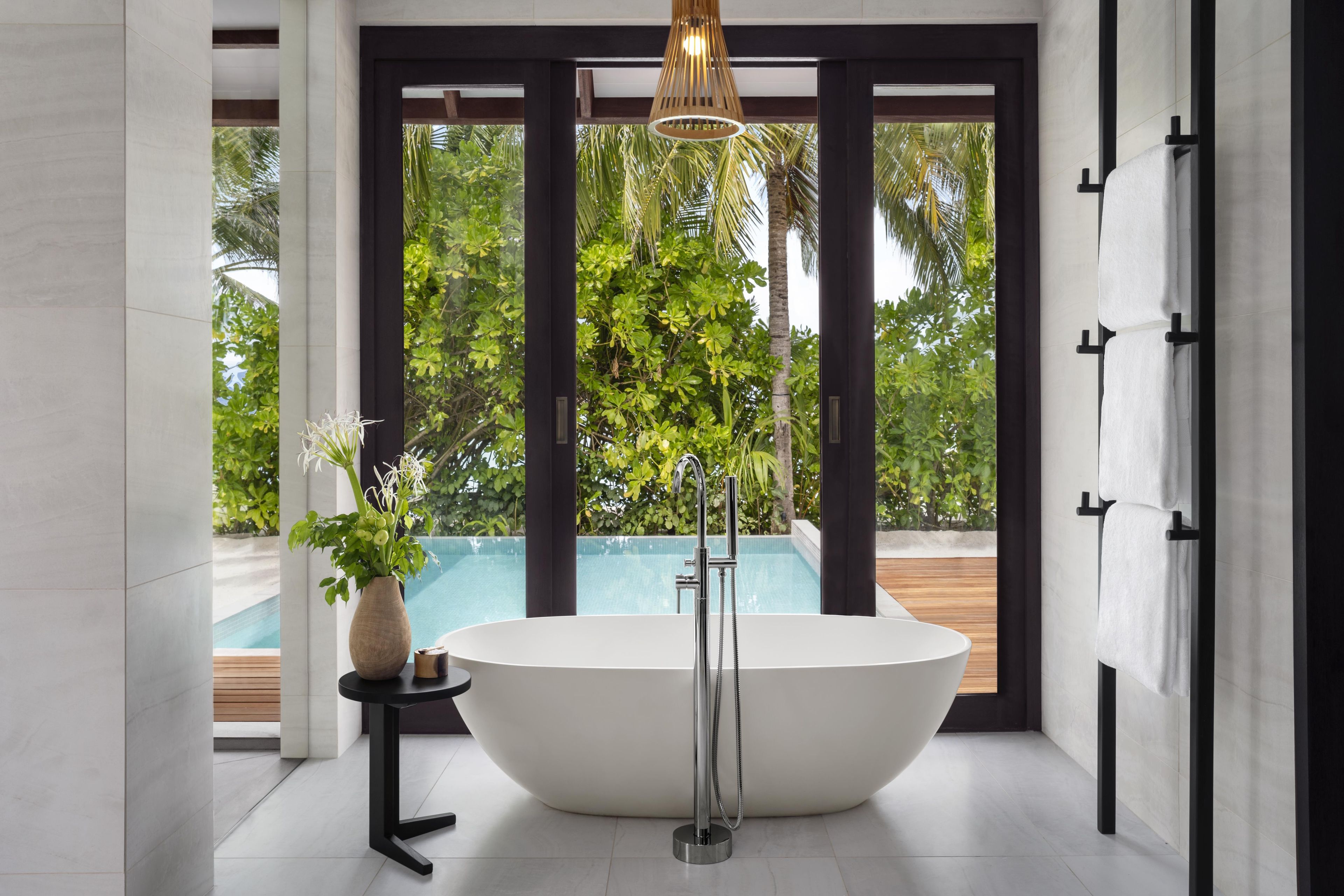 A modern bathroom with a freestanding bathtub, a small table with a vase, and a view of a pool and palm trees.