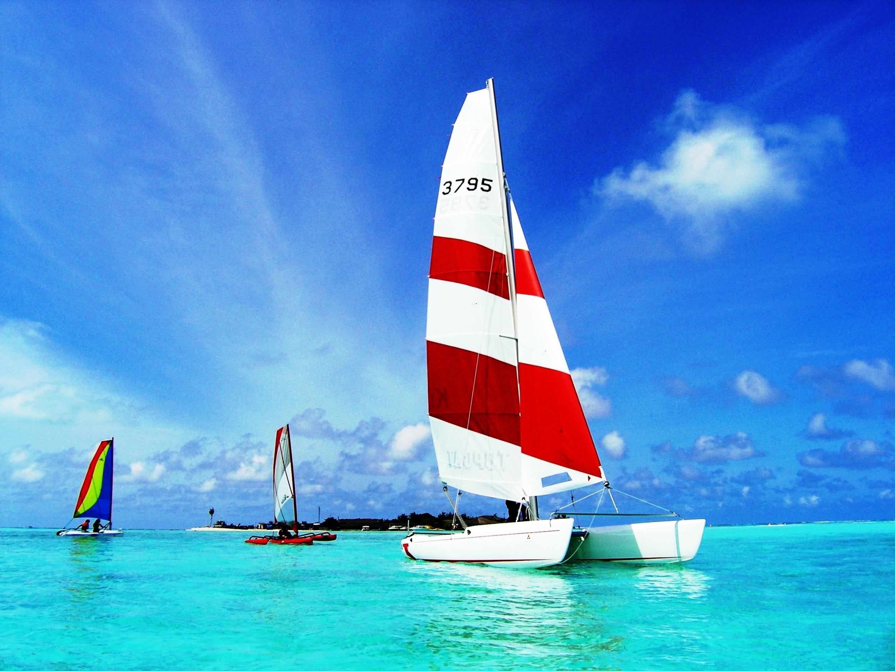 A sailboat with a red and white sail, a smaller sailboat with a multicolored sail, and a distant sailboat with a red sail.