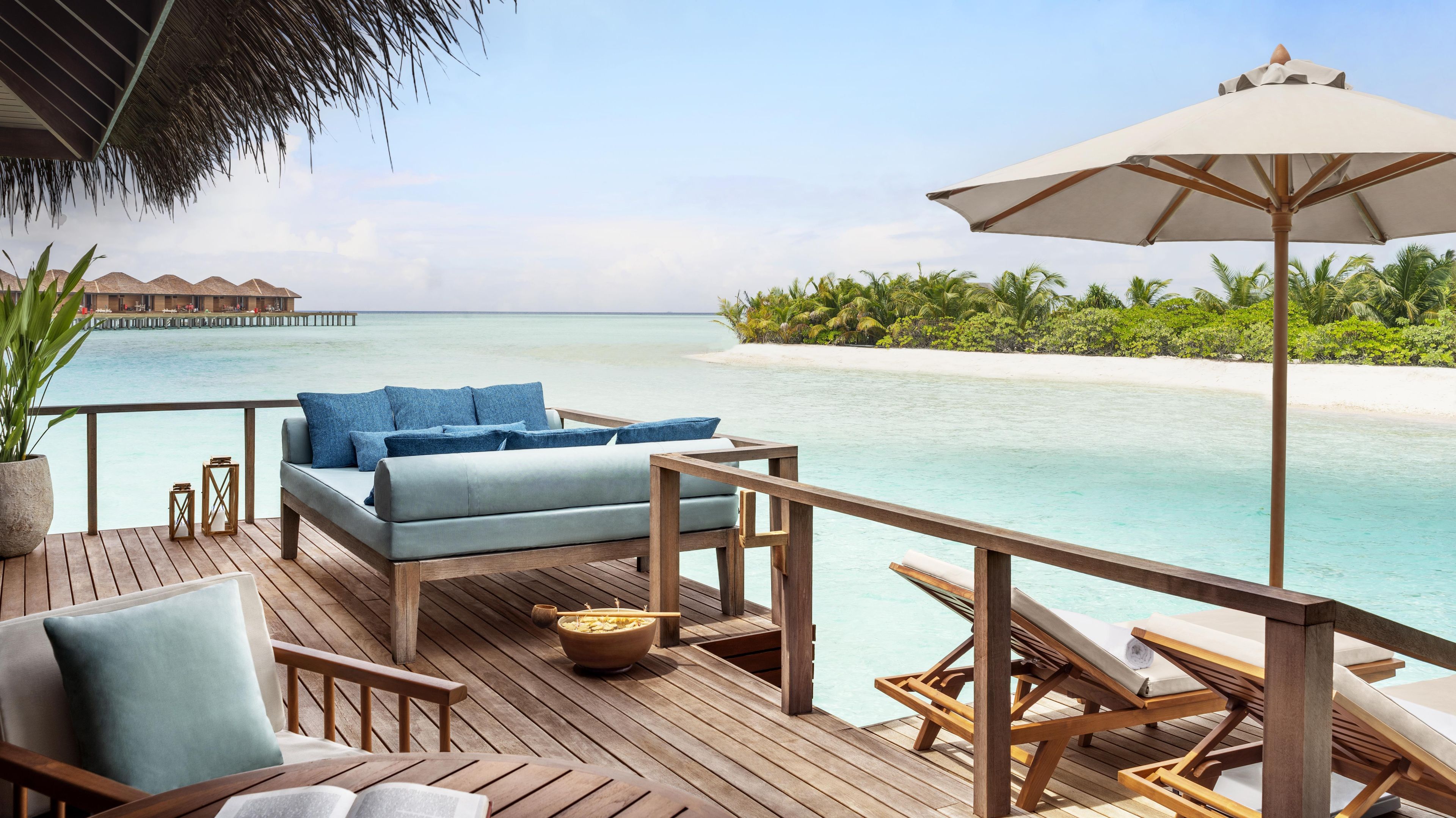 Terraza de madera con sofá y sillones, con vista a una playa de arena blanca y aguas turquesas cristalinas.