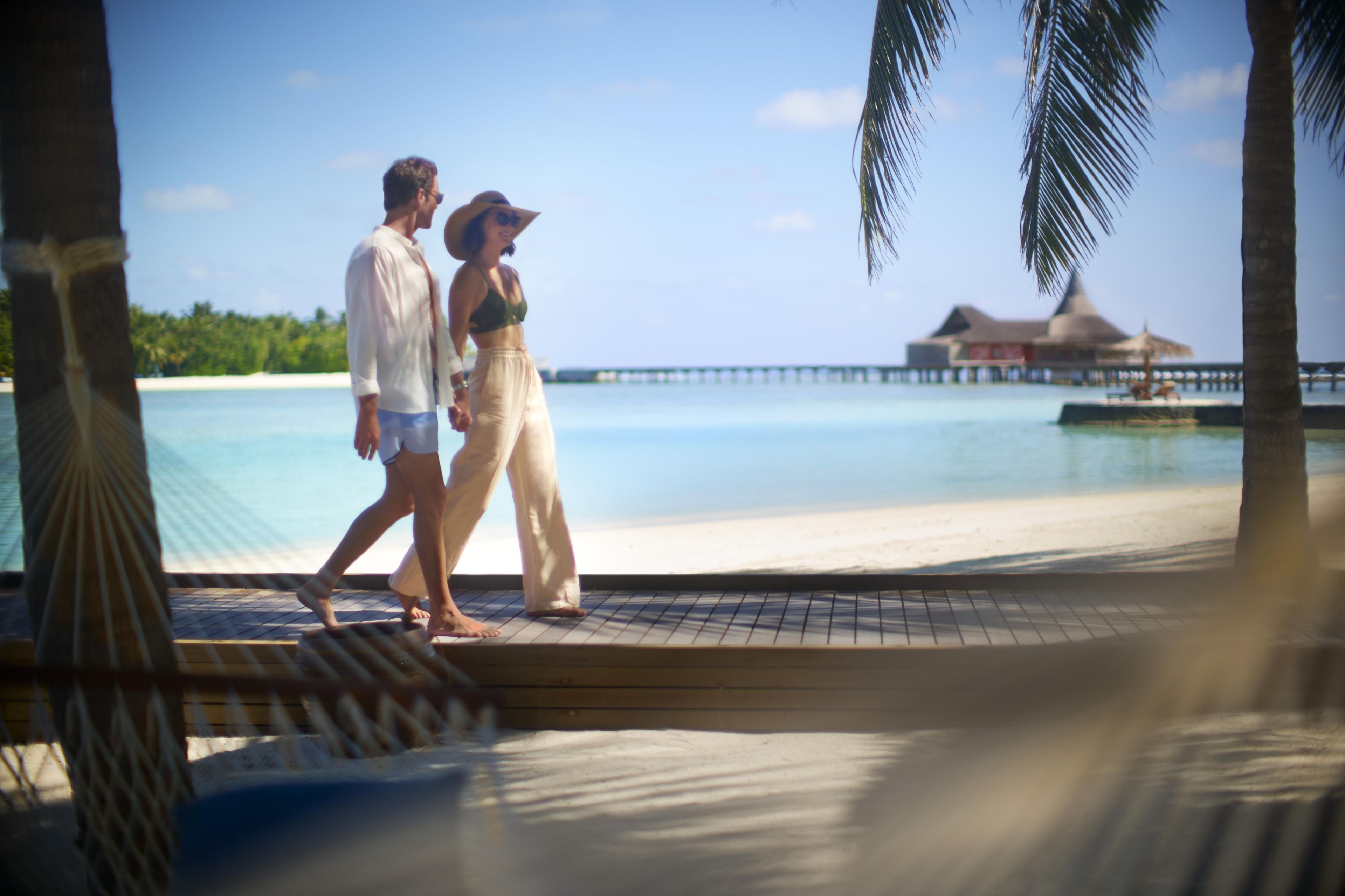 A couple walking on a wooden deck by a beach with a clear view of the ocean and a distant building.