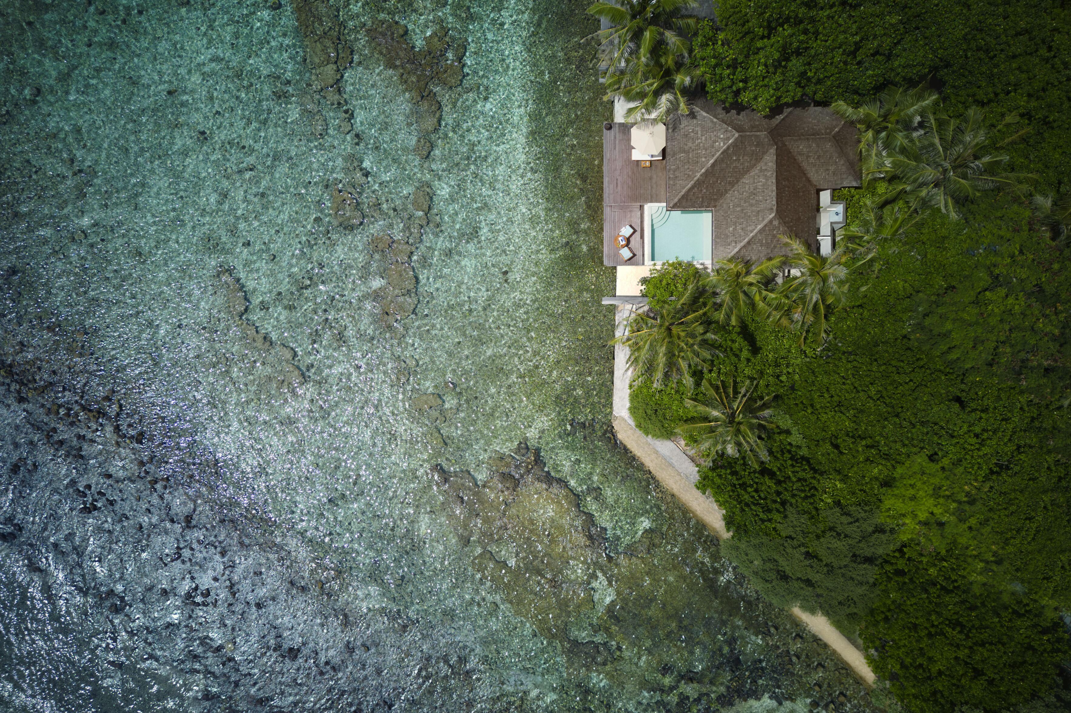 An aerial view of a house with a swimming pool, surrounded by lush greenery and clear turquoise water.
