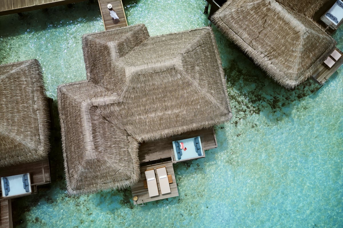Luchtbeeld van bungalows boven het water, met rieten daken, helder turquoise water en houten steigers.