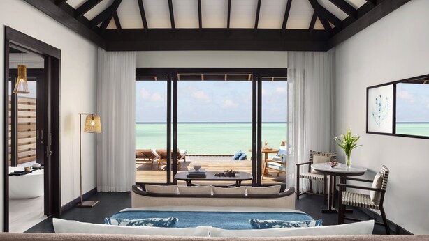 Une chambre d’hôtel dotée d’une grande fenêtre donnant sur la mer, d’un lit, d’un coin salon avec une table et d’un petit balcon.