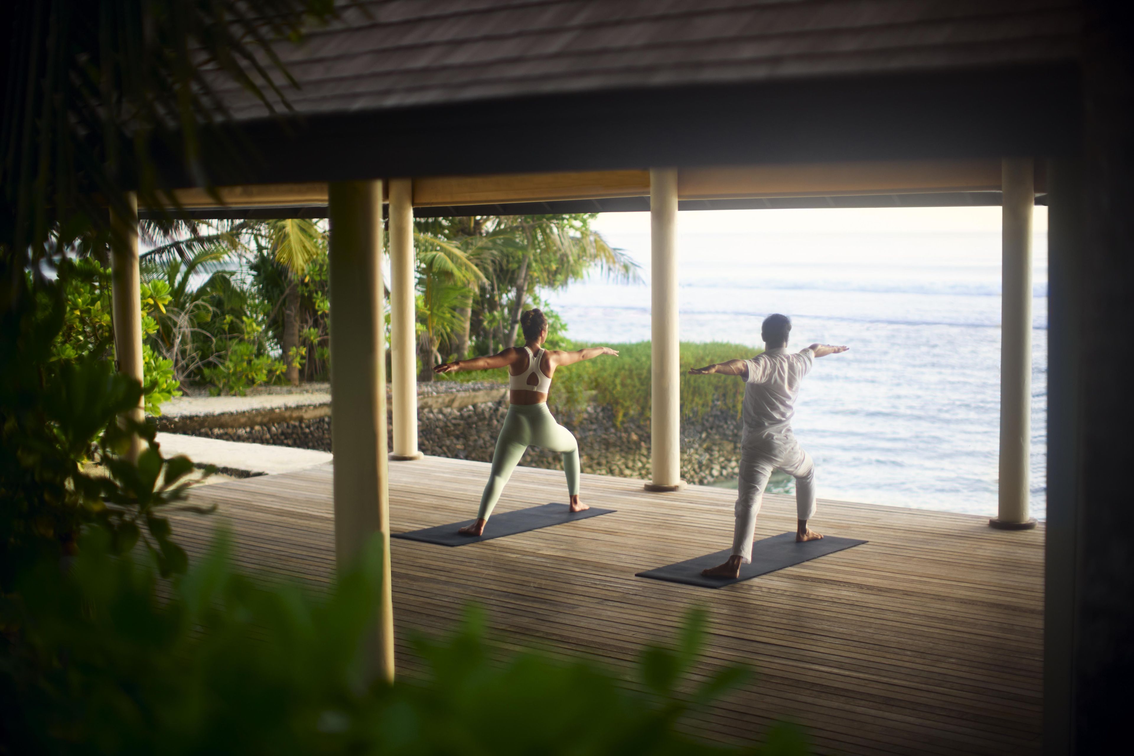 Two people practicing yoga on a wooden deck with a view of the ocean.