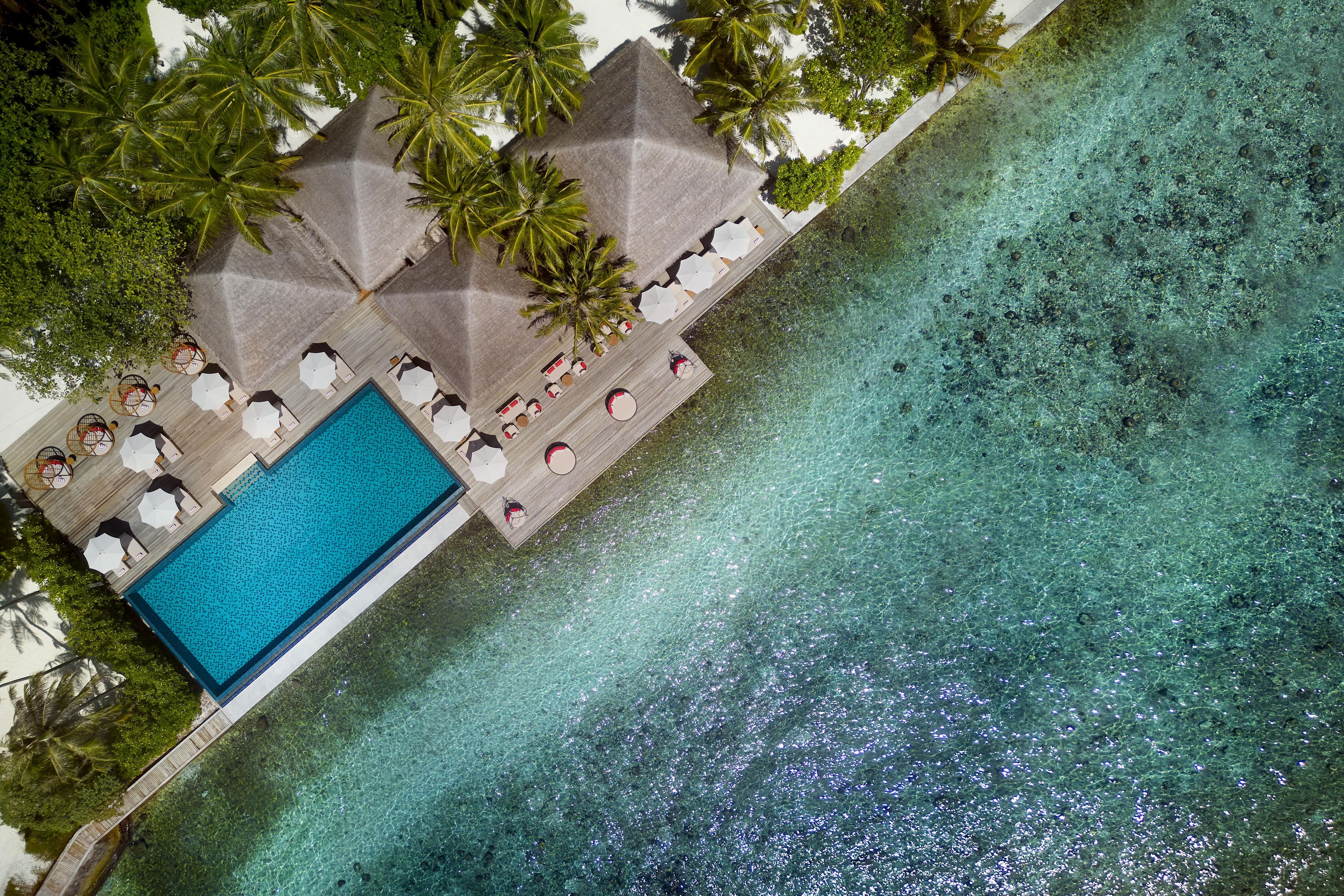 An aerial view of a resort with a clear blue pool, sun loungers, and palm trees.