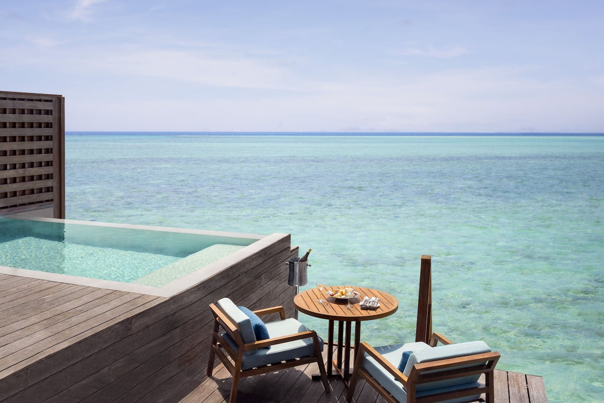 Une terrasse en bois avec une piscine à débordement en verre, des chaises longues et une table avec des boissons et des collations.