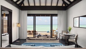 Une chambre d’hôtel dotée d’une grande fenêtre donnant sur l’océan, d’un lit, d’un coin salon avec une table et d’un petit balcon.