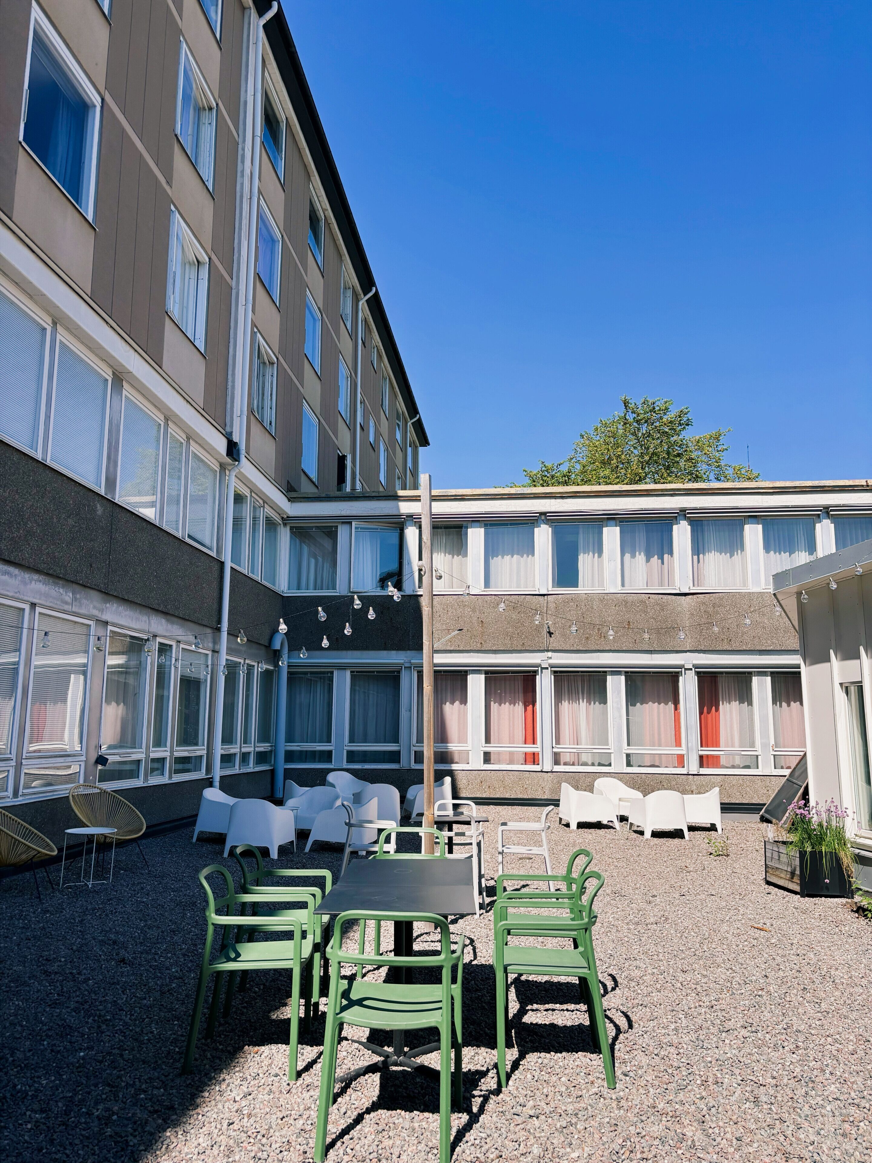 An outdoor seating area with tables and chairs, a gravel ground, and a building with multiple windows and balconies.