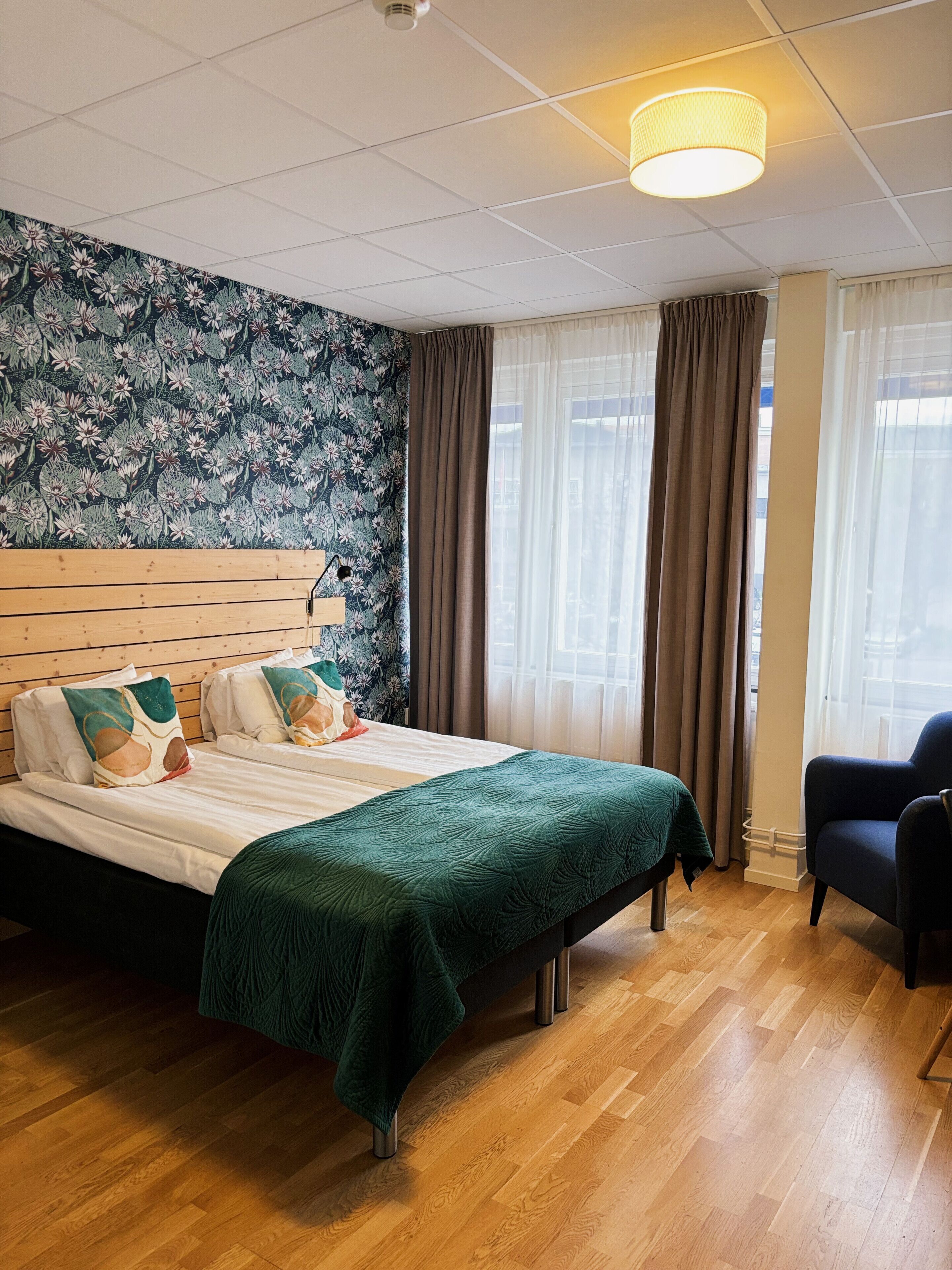 A hotel room with a large bed, a wooden headboard, a green bedspread, a blue armchair, and a floral wallpaper.