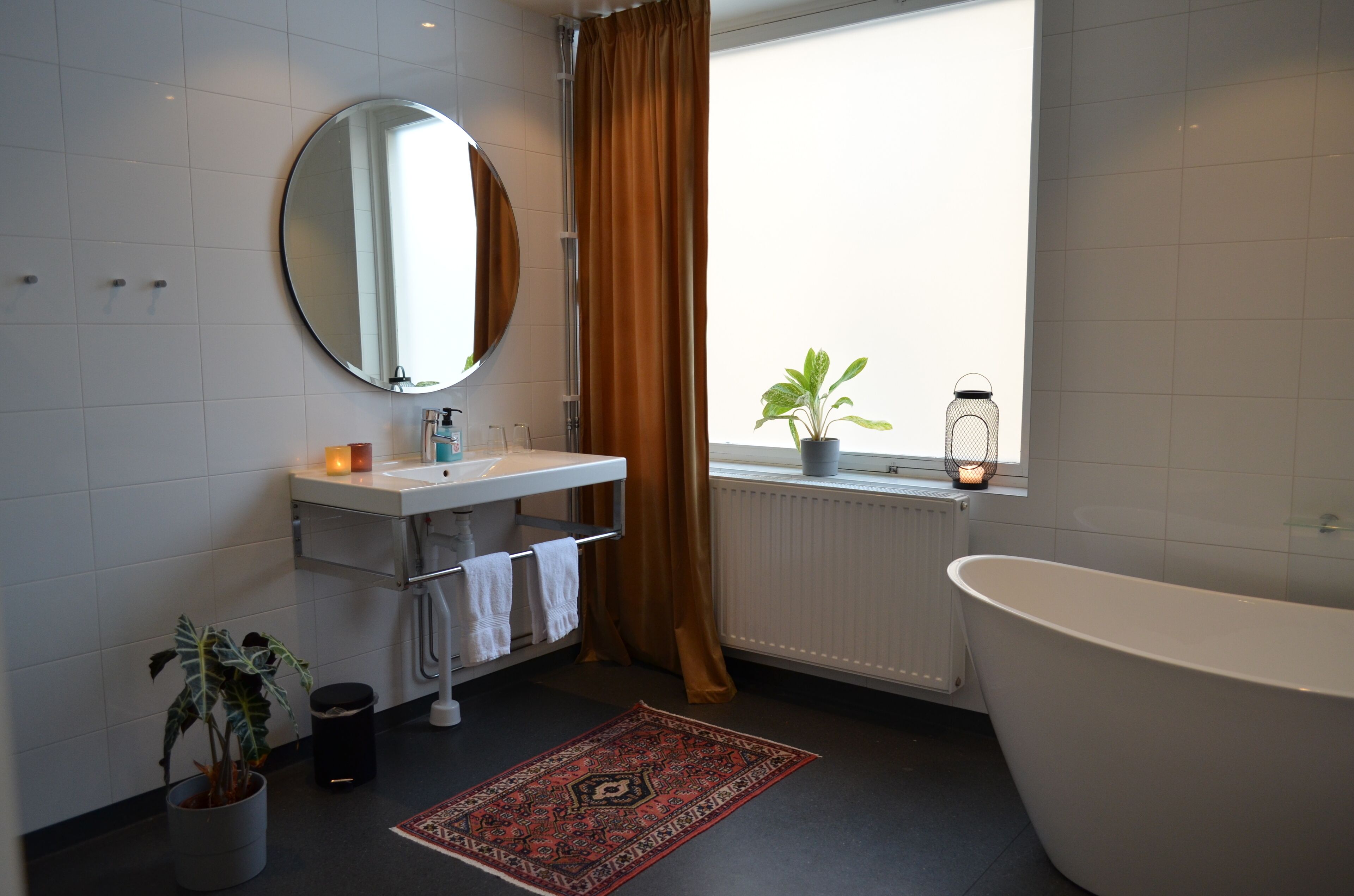 Ett modernt badrum med ett fristående badkar, en rund spegel, en tvättställ med handduksställ och ett fönster med gardiner.