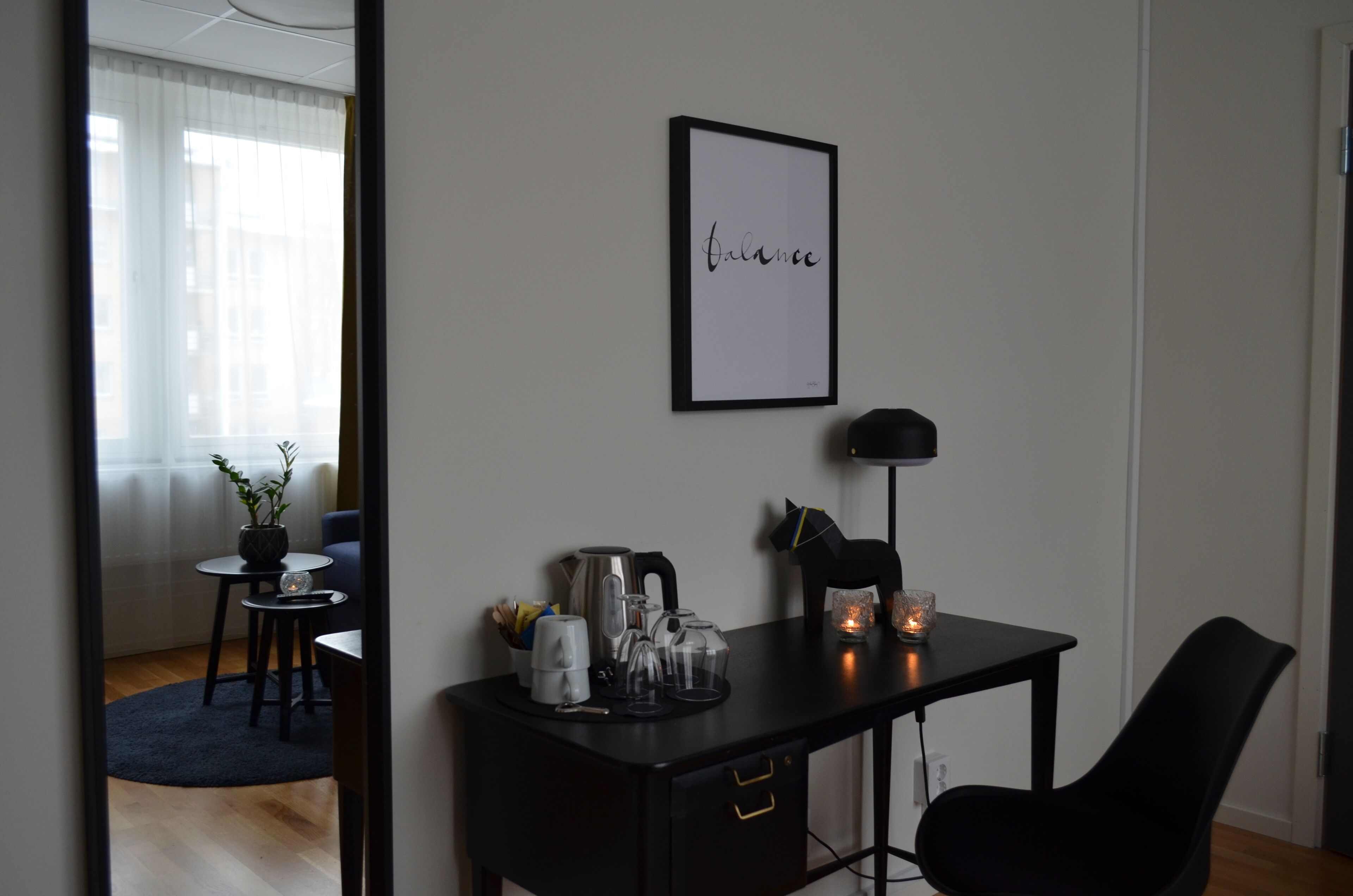 Una habitación moderna y minimalista con un escritorio negro, una obra de arte enmarcada con la palabra 'balance', una planta en maceta y una pequeña mesa con cafetera y tazas.