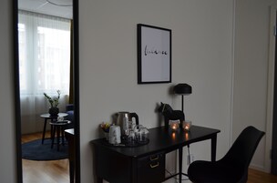 Una habitación moderna y minimalista con un escritorio negro, una obra de arte enmarcada con la palabra 'balance', una planta en maceta y una pequeña mesa con cafetera y tazas.