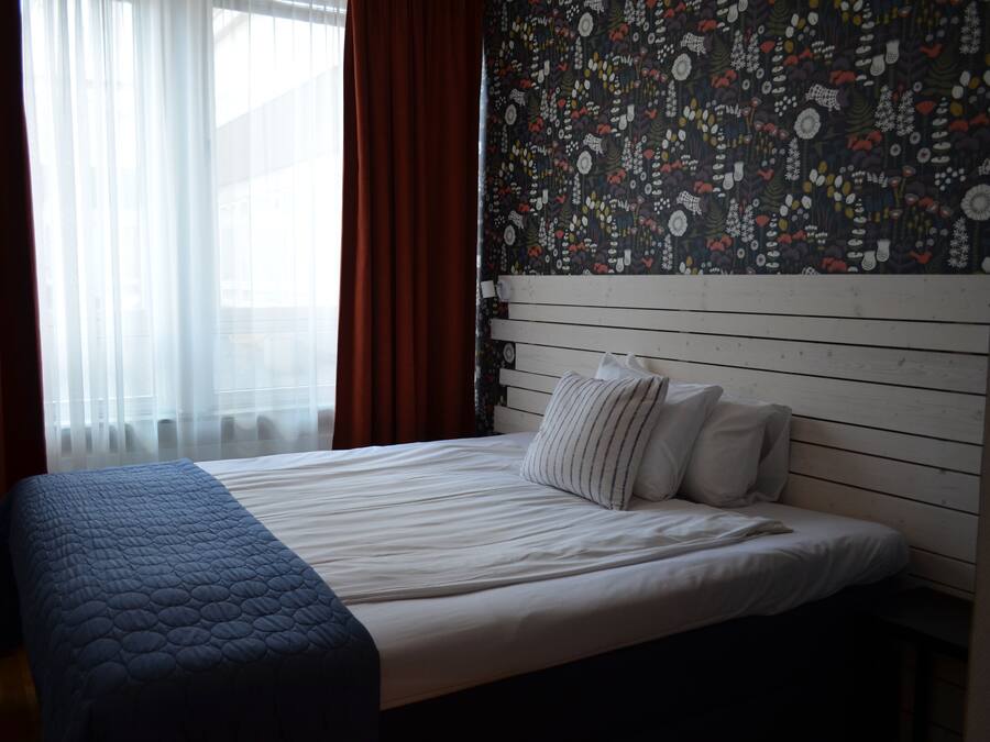 Una camera d'albergo con un letto, una finestra con tende leggere, un muro decorato e una testiera in legno.