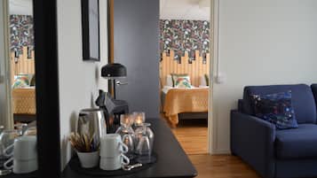 Una habitación con una cama, un sofá azul, una mesa con tazas y vasos, y un espejo que refleja la habitación.