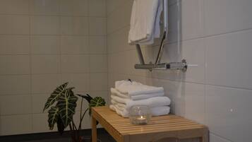 A bathroom with white tiled walls, a wooden bench, a stack of white towels, a glass candle holder, and a potted plant.