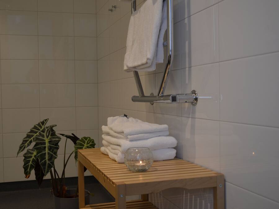 A bathroom with white tiled walls, a wooden bench, a stack of white towels, a glass candle holder, and a potted plant.