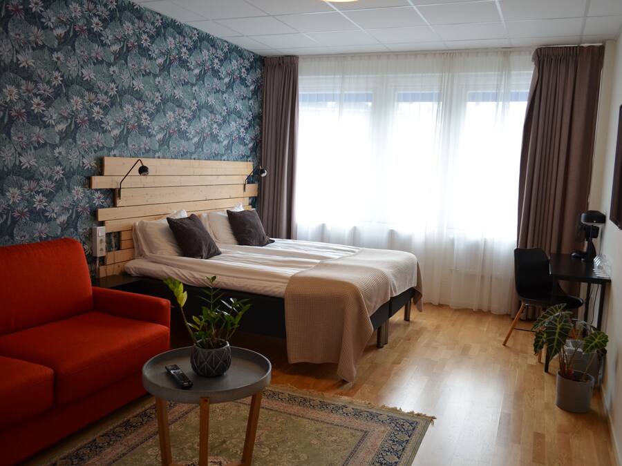A hotel room with a wooden headboard bed, a red sofa, a small table with a plant, and a desk with a chair.
