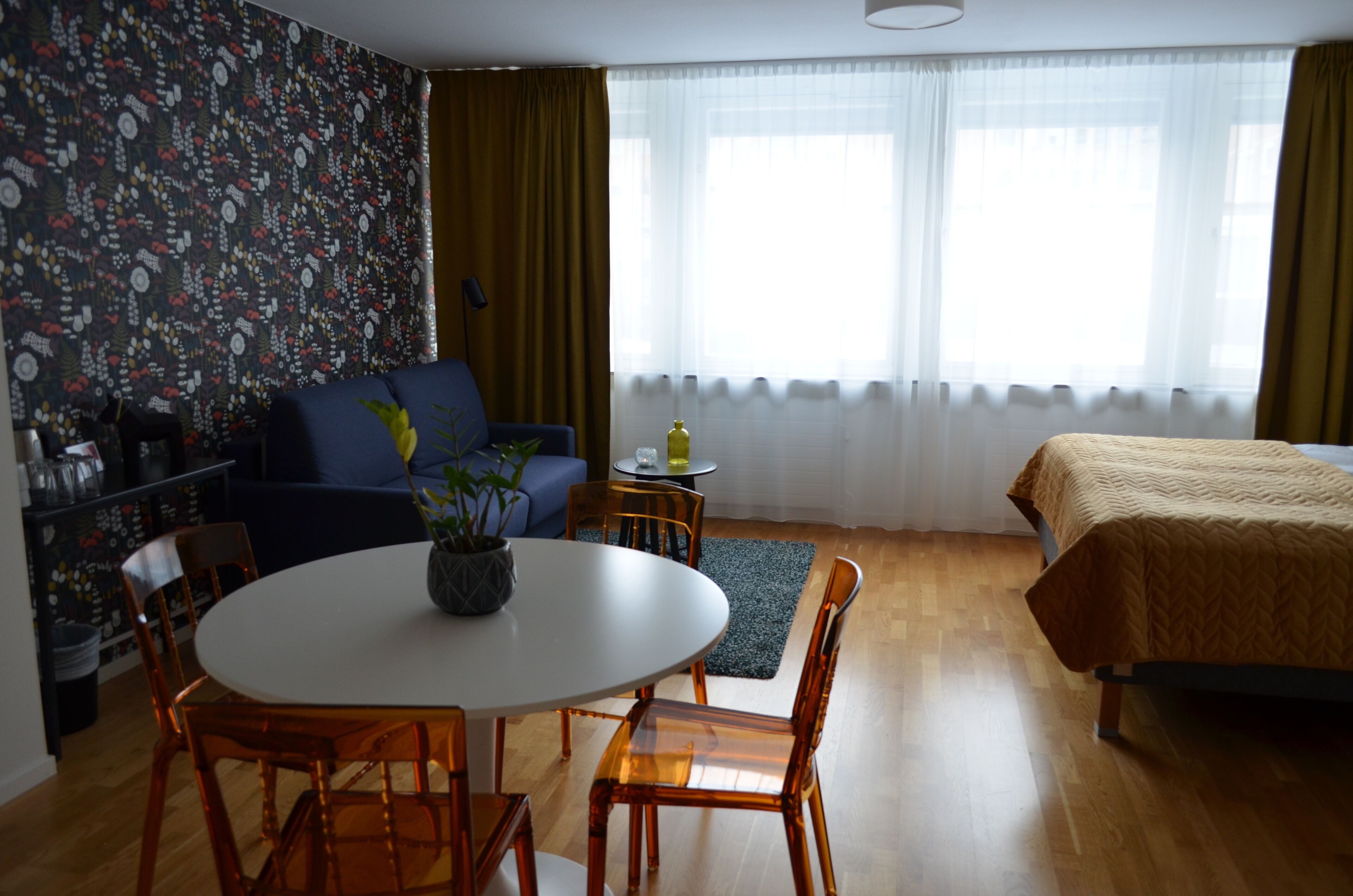 Una habitación con una cama, un sofá, una mesa de comedor con sillas, una mesita auxiliar y una ventana con cortinas.