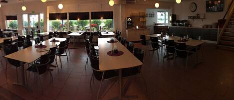 A spacious dining area with multiple tables and chairs, large windows with views of greenery, and a kitchen area in the background.