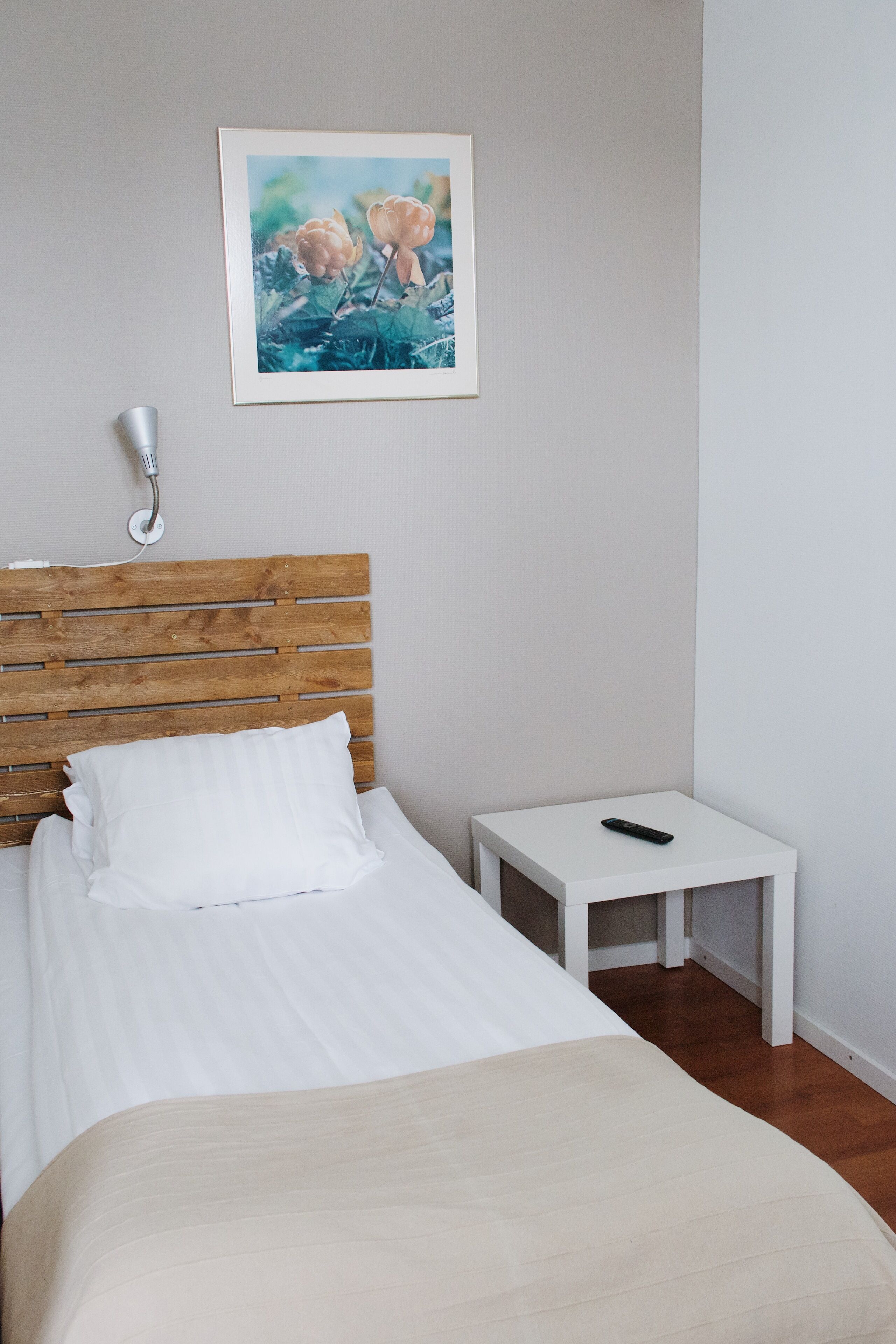 A neatly made bed with white linens, a wooden headboard, a bedside table with a remote control, and a framed picture on the wall.