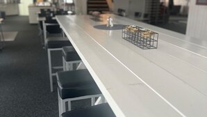 Una barra larga y blanca con taburetes negros, iluminación colgante y una escalera al fondo.