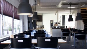 Un comedor moderno con sillas y mesas negras, iluminación colgante y una cocina al fondo.