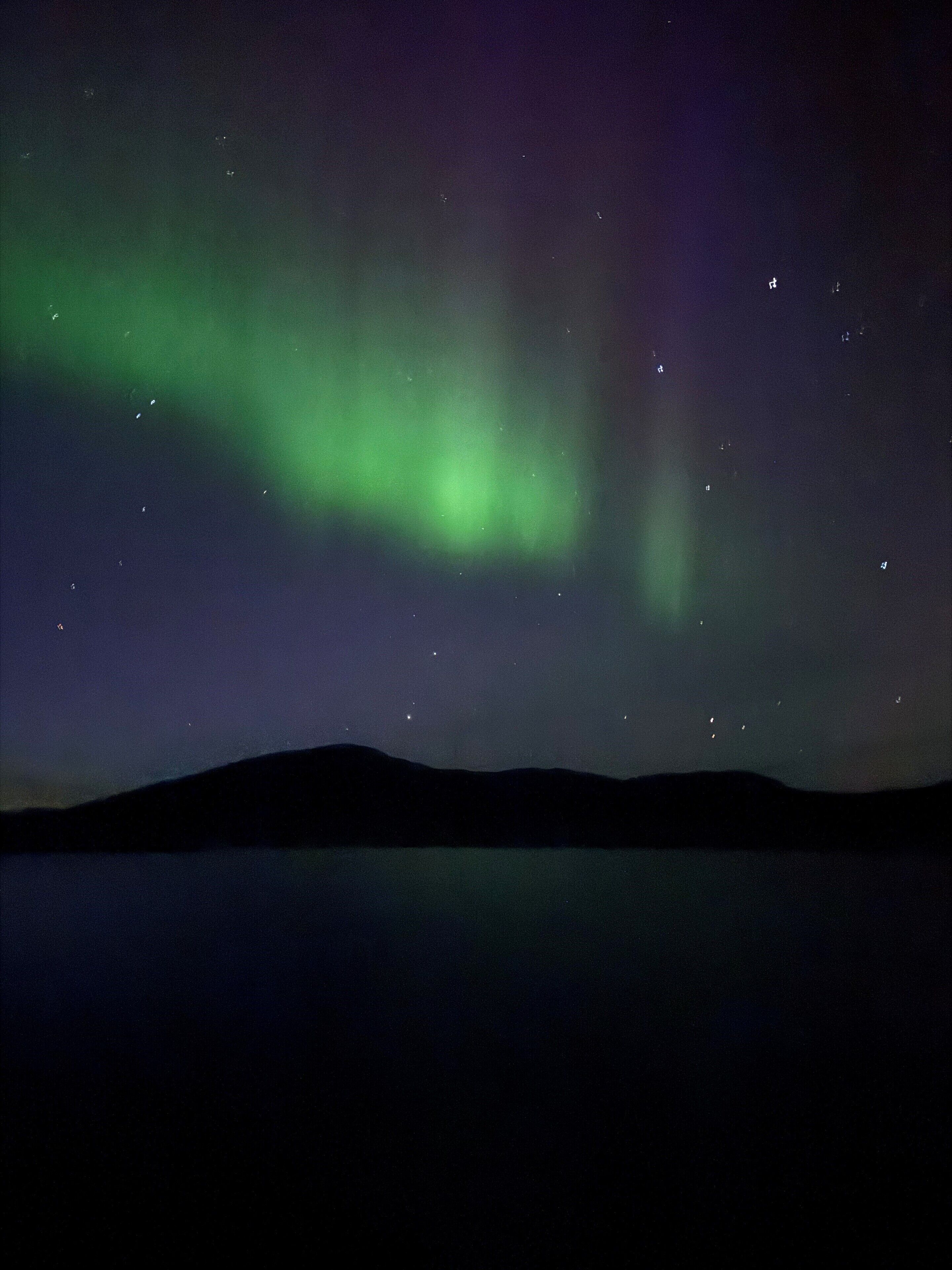 A night sky with the Northern Lights over a calm body of water and a silhouette of a mountain range.