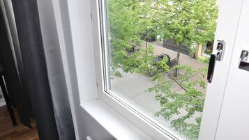 Ein Zimmer mit Fensterblick auf eine Straße mit Bäumen und Außenbestuhlung.