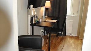 A compact room with a wooden floor, a black leather chair, a small table with a lamp, a television on a stand, and a window with curtains.