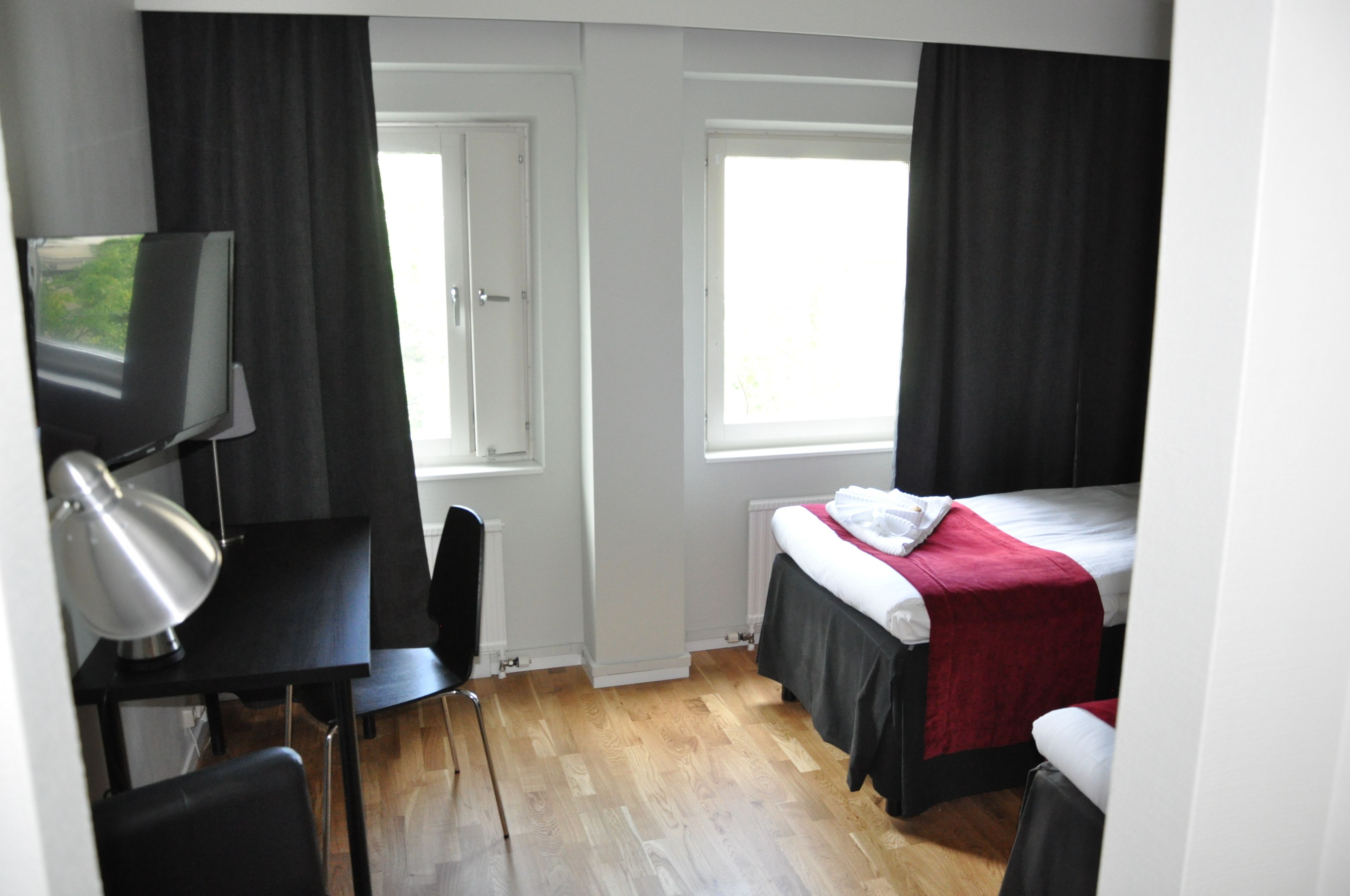 Una habitación pequeña y limpia con cama, escritorio, silla, televisor y dos ventanas.