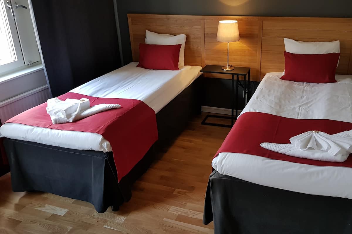 Two single beds with red and white bedding, a wooden headboard, a bedside table with a lamp, and a window with curtains.