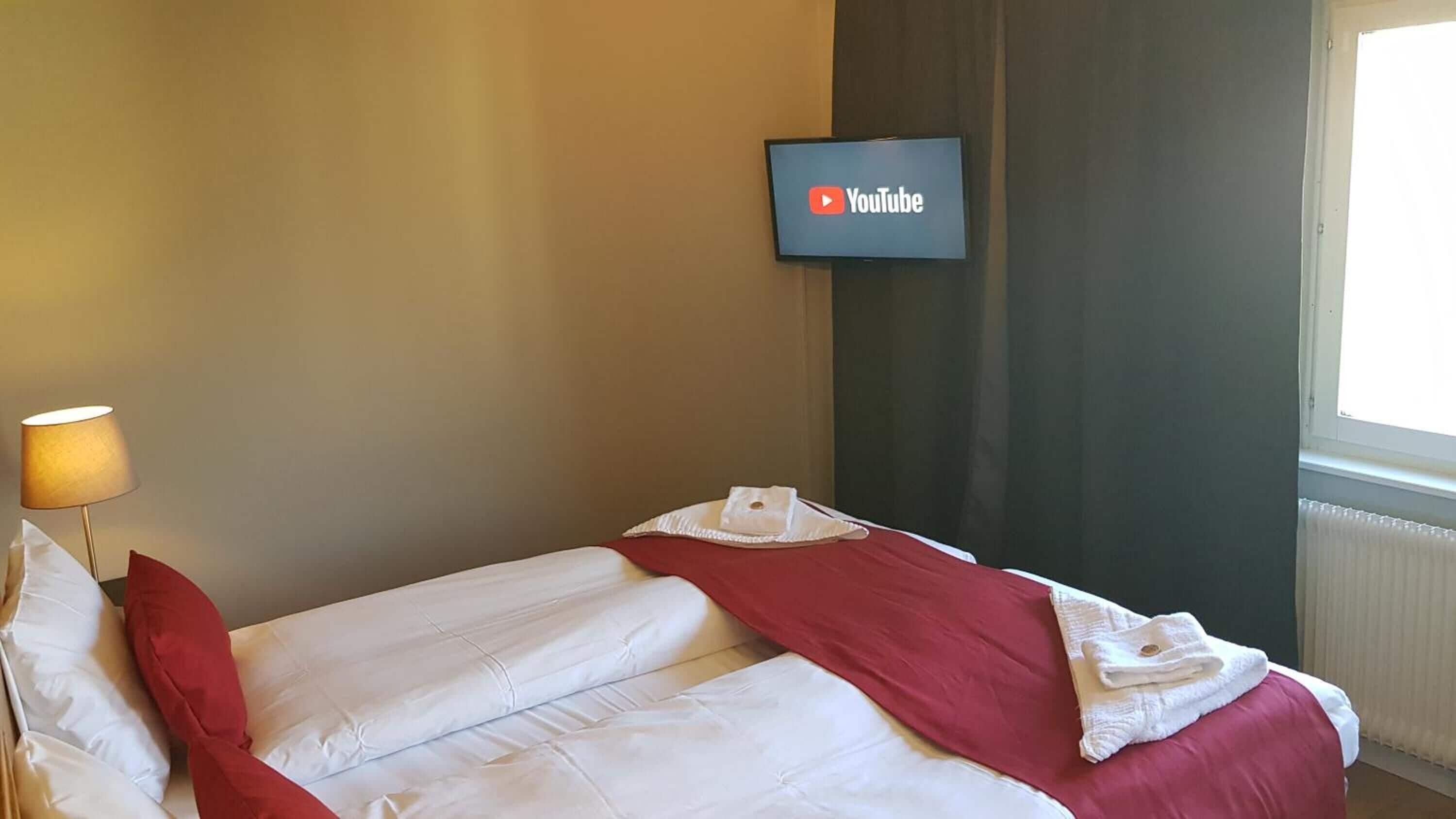 A hotel room with a bed, a TV mounted on the wall displaying the YouTube logo, a lamp, and a window with curtains.