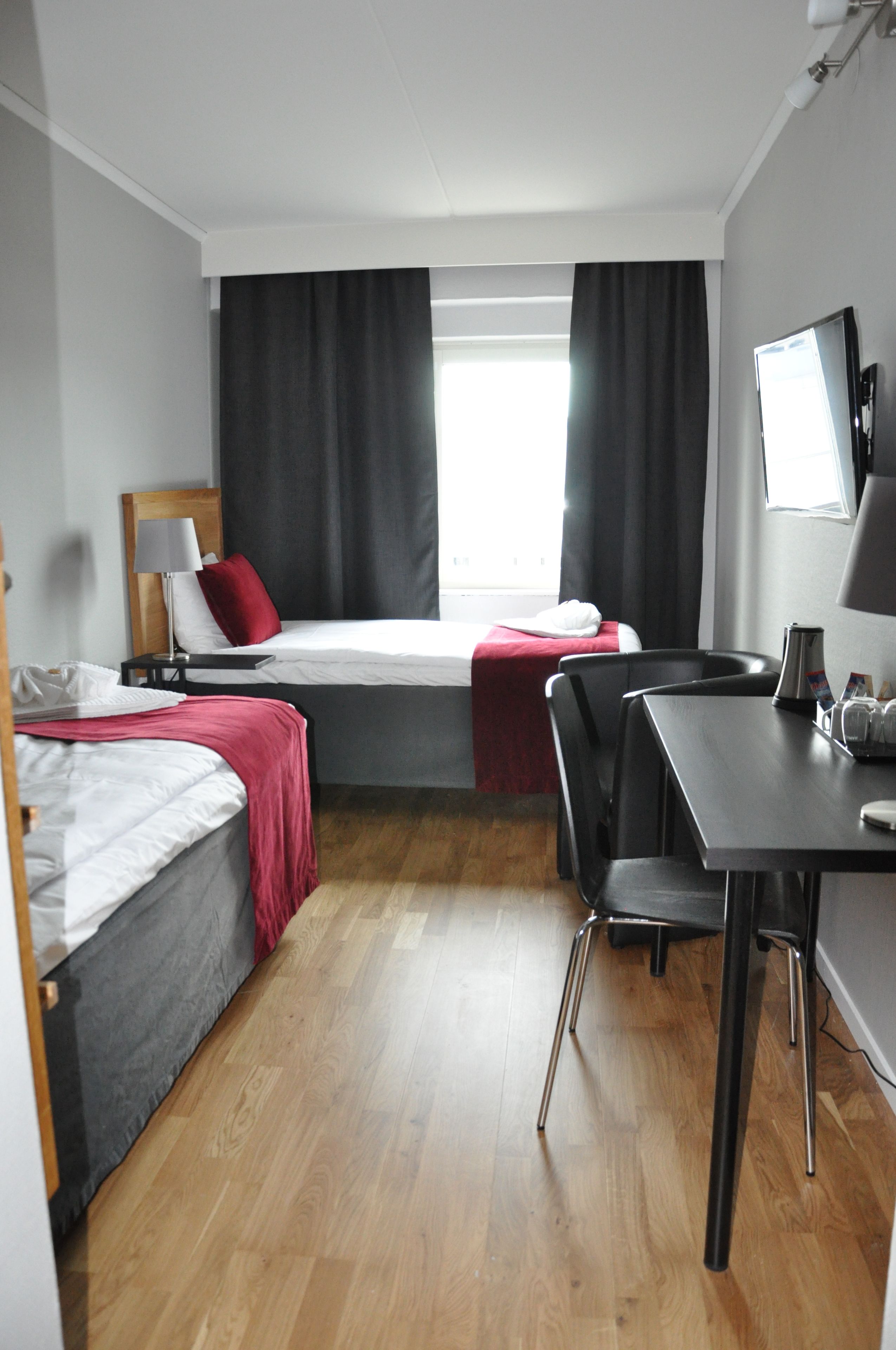 A compact hotel room with two single beds, a small desk, a chair, a TV, and a window with curtains.