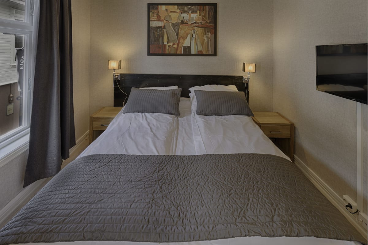 A hotel room with a large bed, two bedside tables with lamps, a painting on the wall, and a TV mounted on the wall.