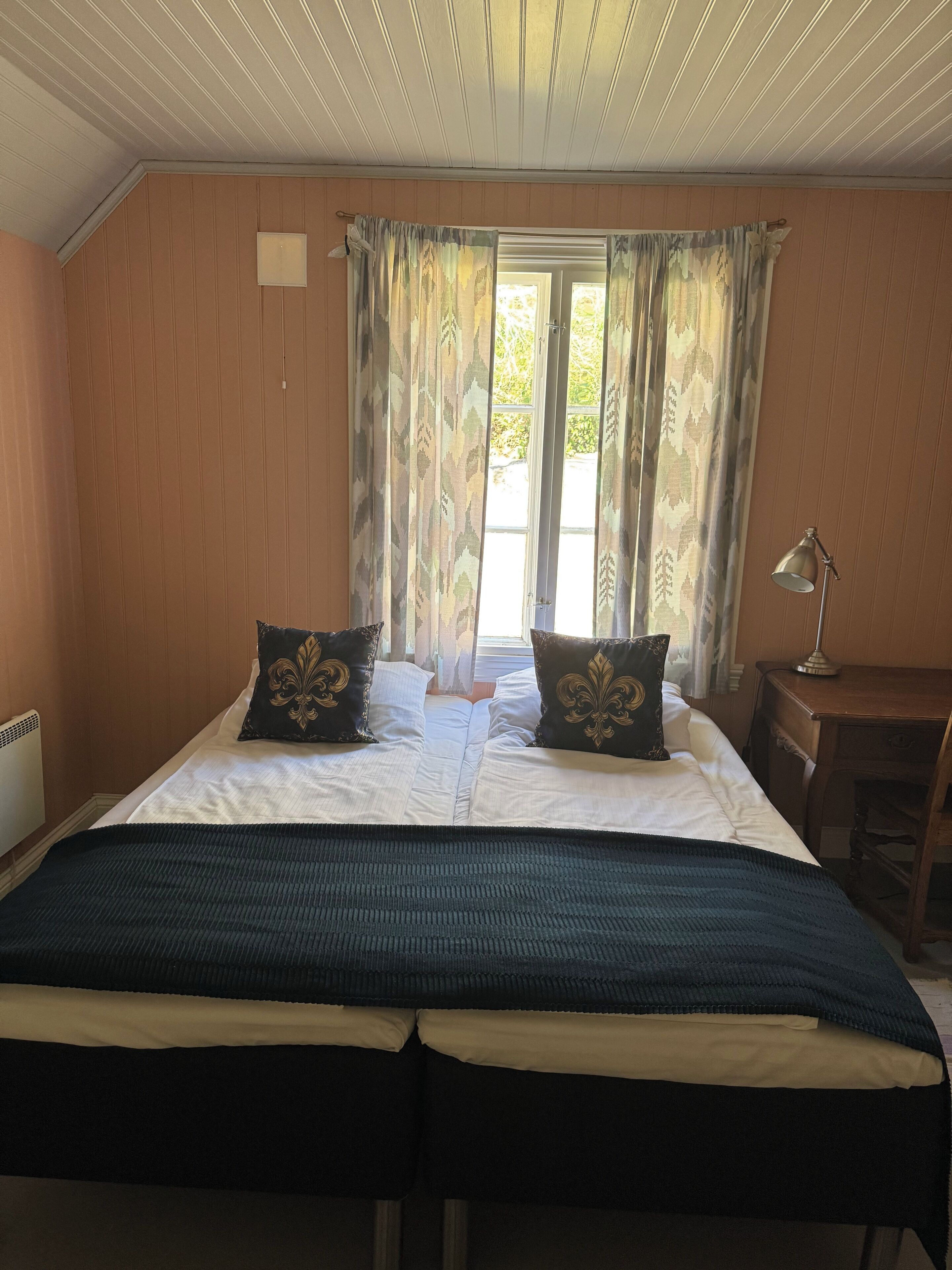A room with a bed, two pillows, a desk, a lamp, and a window with curtains.