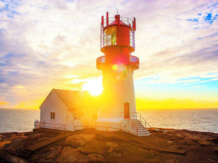 Un faro de exterior blanco con detalles rojos, situado en una costa rocosa con vista al mar.