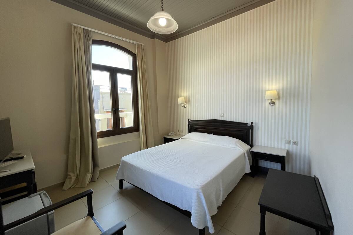 Habitación de hotel con cama, mesitas de noche, escritorio con silla, ventana con cortinas e iluminación en el techo.