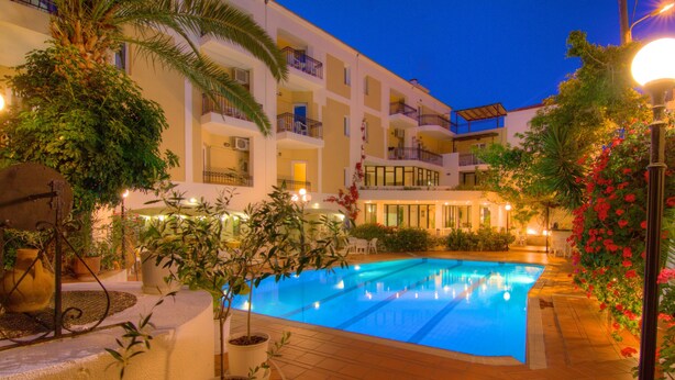 Una piscina rodeada de plantas en macetas y muebles de exterior, con un edificio al fondo.