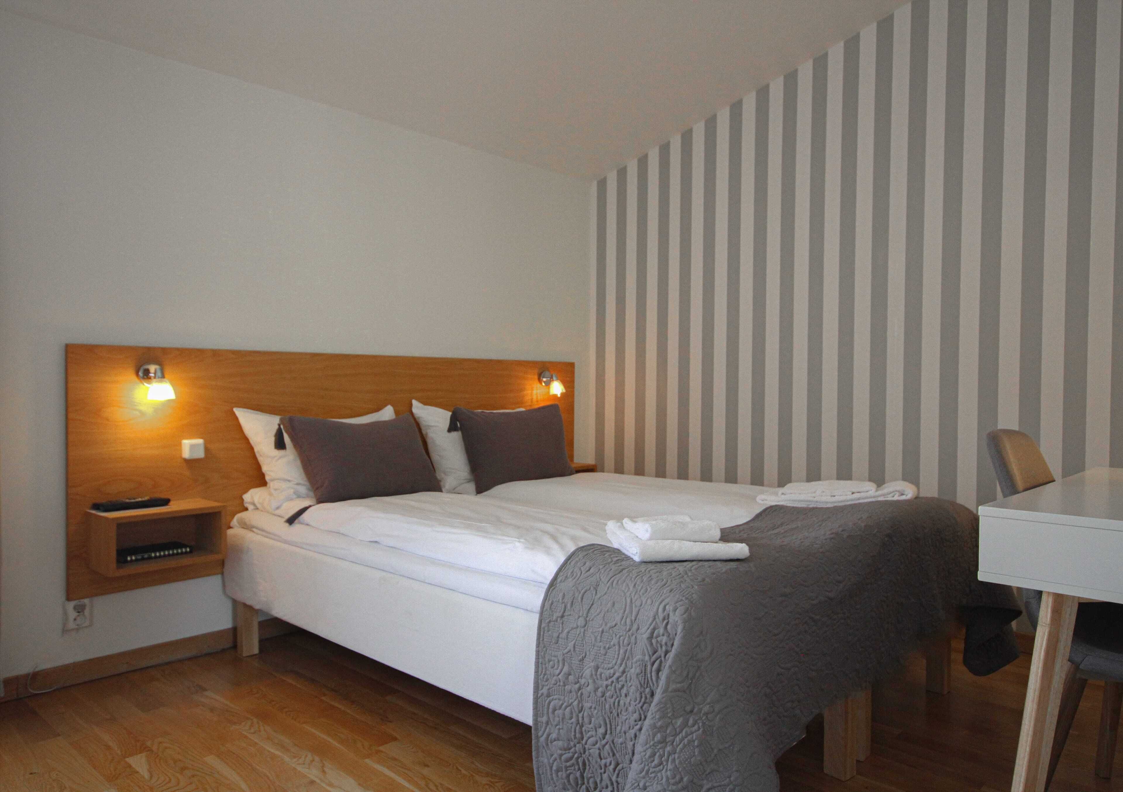 A neatly made bed with a wooden headboard, a bedside table with a lamp, and a striped wall in the background.