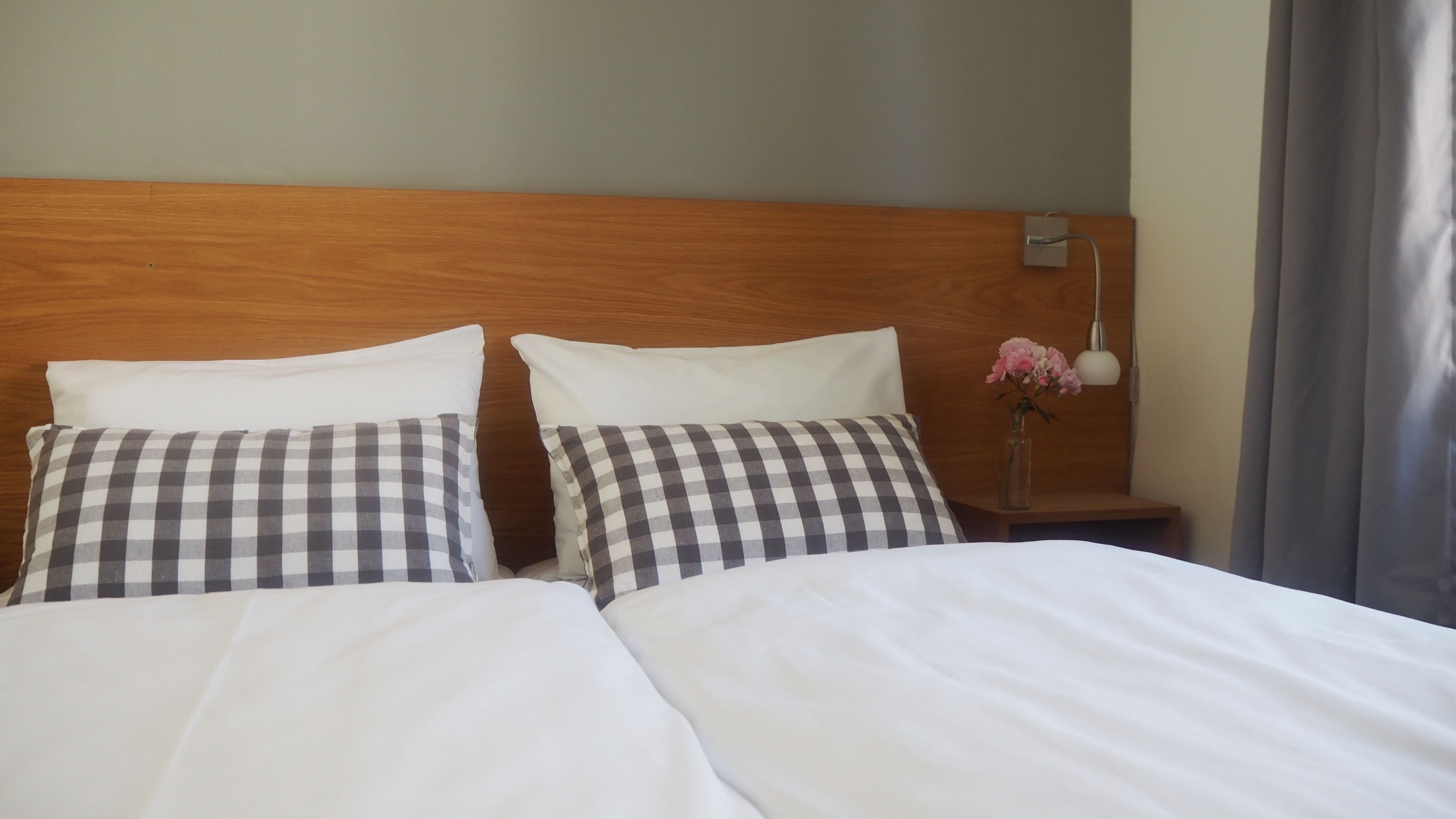 A hotel room with two beds, checkered pillows, a wooden headboard, a nightstand with a vase of flowers, and a lamp.