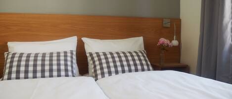 A hotel room with two beds, checkered pillows, a wooden headboard, a nightstand with a vase of flowers, and a lamp.