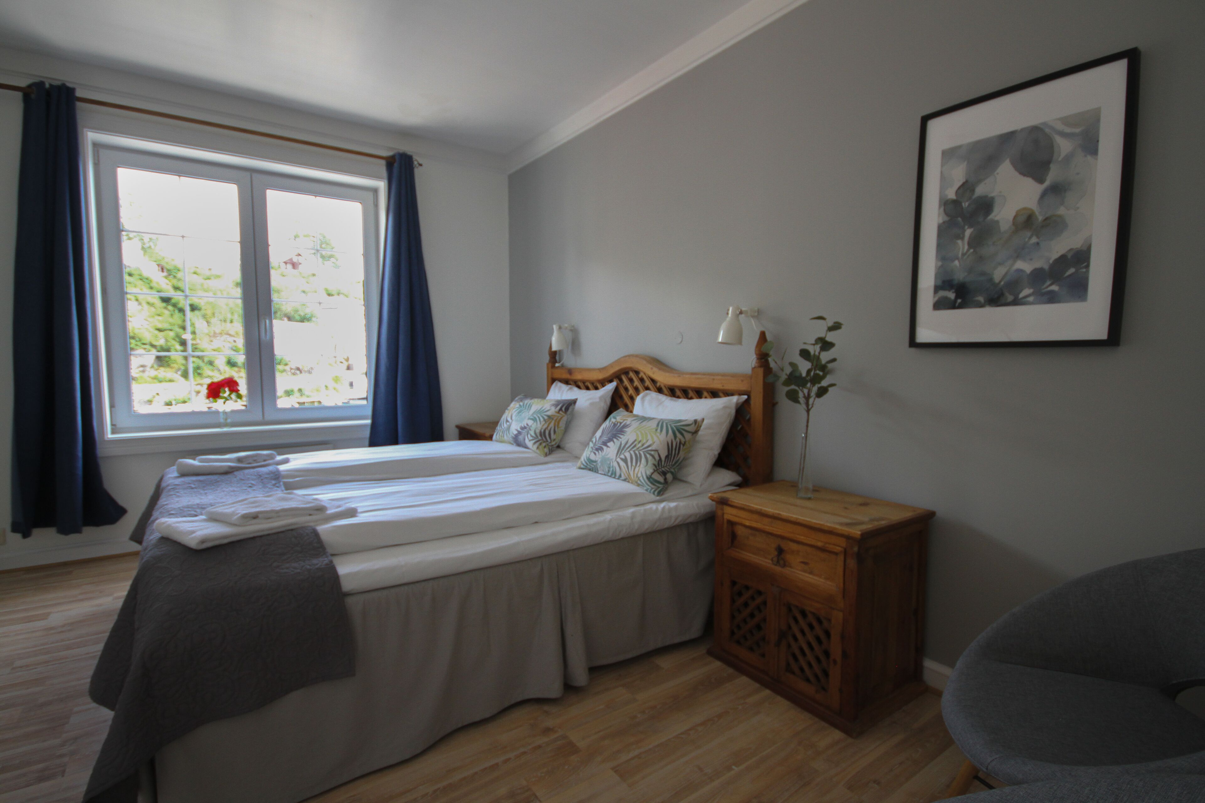 Una habitación con dos camas individuales, una mesita de noche de madera, una silla, una ventana con cortinas y un cuadro en la pared.