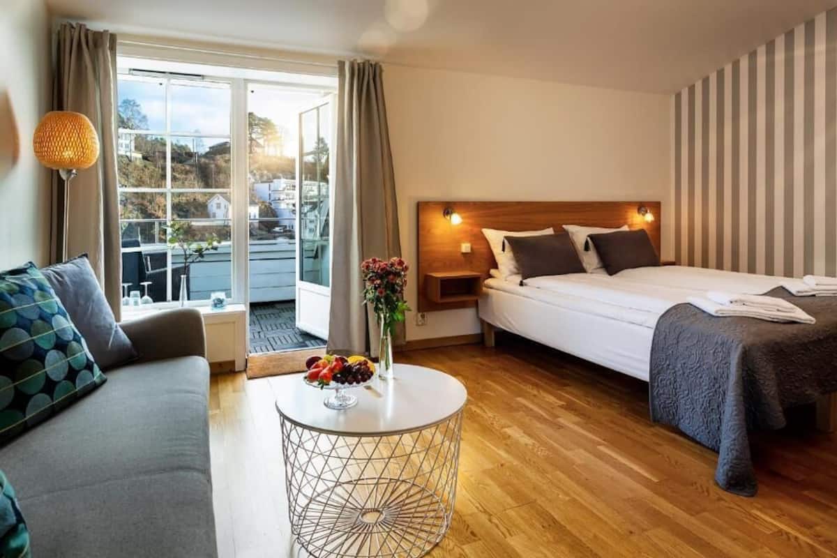 A hotel room with a bed, a sofa, a round table with fruit, and a view of the outside.