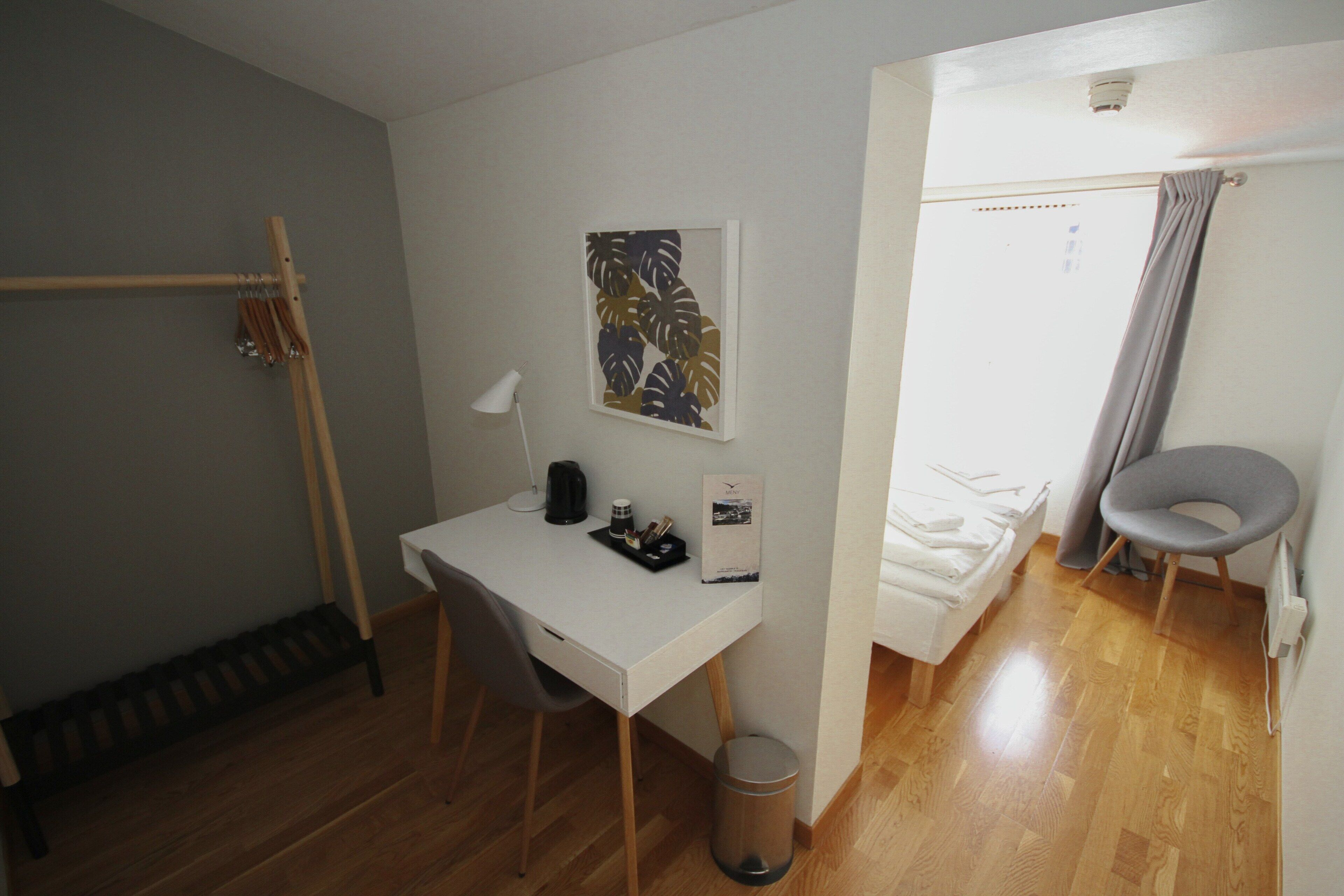 A compact room with a white desk, a chair, a small trash bin, a bed with a grey chair, and a wall-mounted picture.